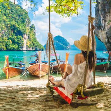 Woman on a swing overlooking a serene beach with traditional longtail boats included in our Thailand tour packages.